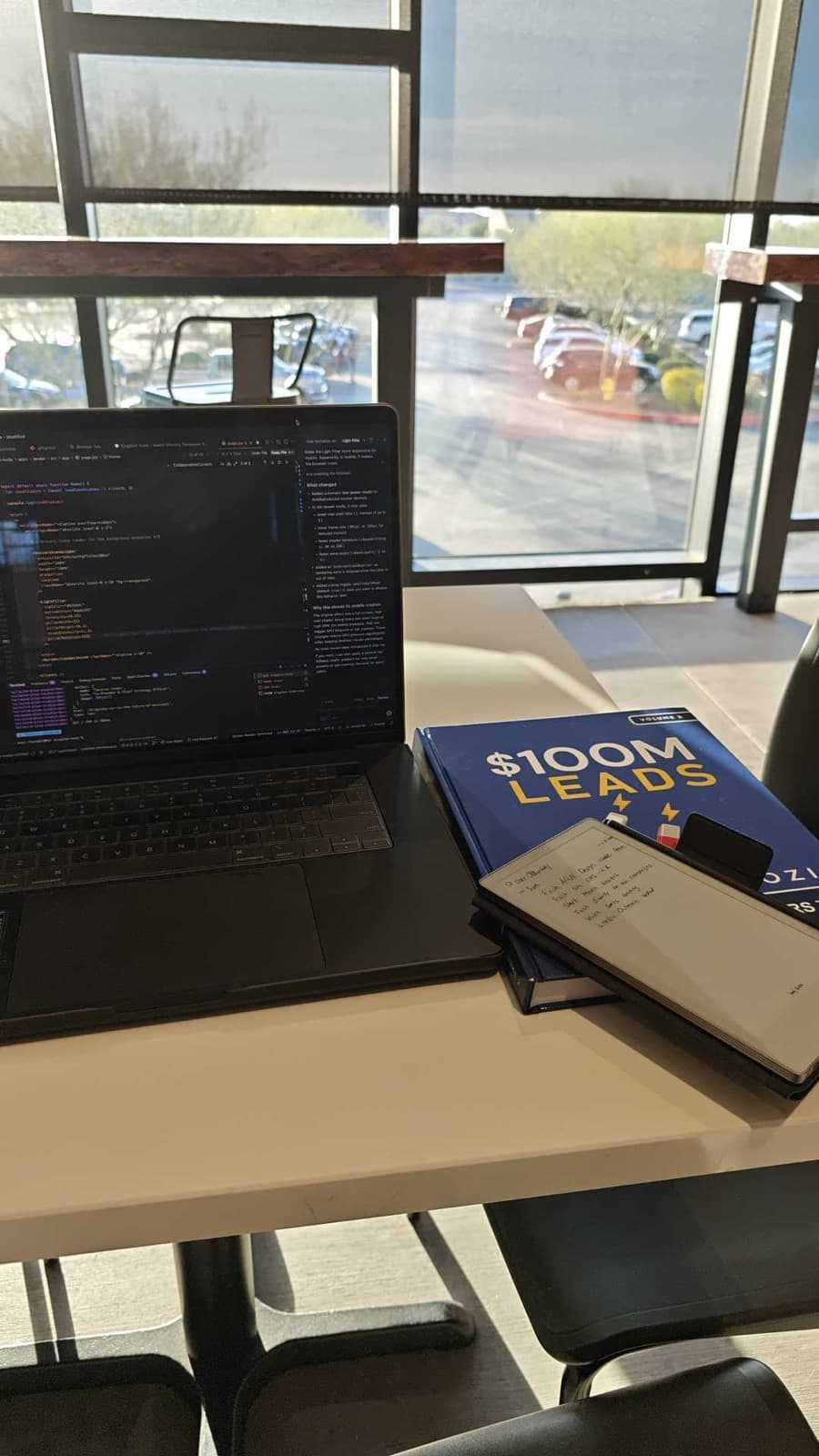 A laptop running code beside a copy of $100M Leads and a notebook, in the window light of a co-working space